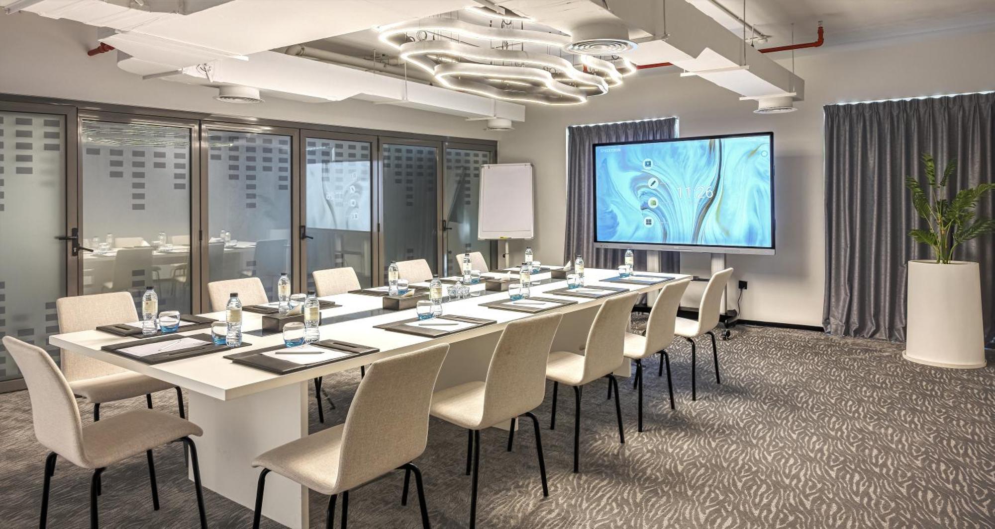 Millennium Atria Business Bay Aparthotel Dubaj Kültér fotó The photo shows a modern conference room set up for a meeting. There is a long table with several chairs arranged around it, each with a notepad and pen. A large screen displays a graphic or presentation, and there is a whiteboard in the corner. The 