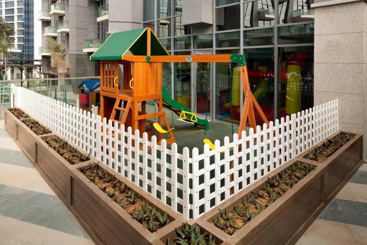 Millennium Atria Business Bay Aparthotel Dubaj Kültér fotó The image shows a children's playground area, which is enclosed by a white picket fence. The playground features a wooden play structure with a slide, swings, and climbing elements. Surrounding the play area are planters with greenery, and the backdr
