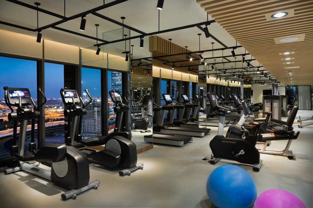 Millennium Atria Business Bay Aparthotel Dubaj Kültér fotó The photo shows a modern gym with various types of exercise equipment available. There are several treadmills and elliptical machines arranged in rows, with large windows in the background providing a view of the city. The lighting is bright and cont