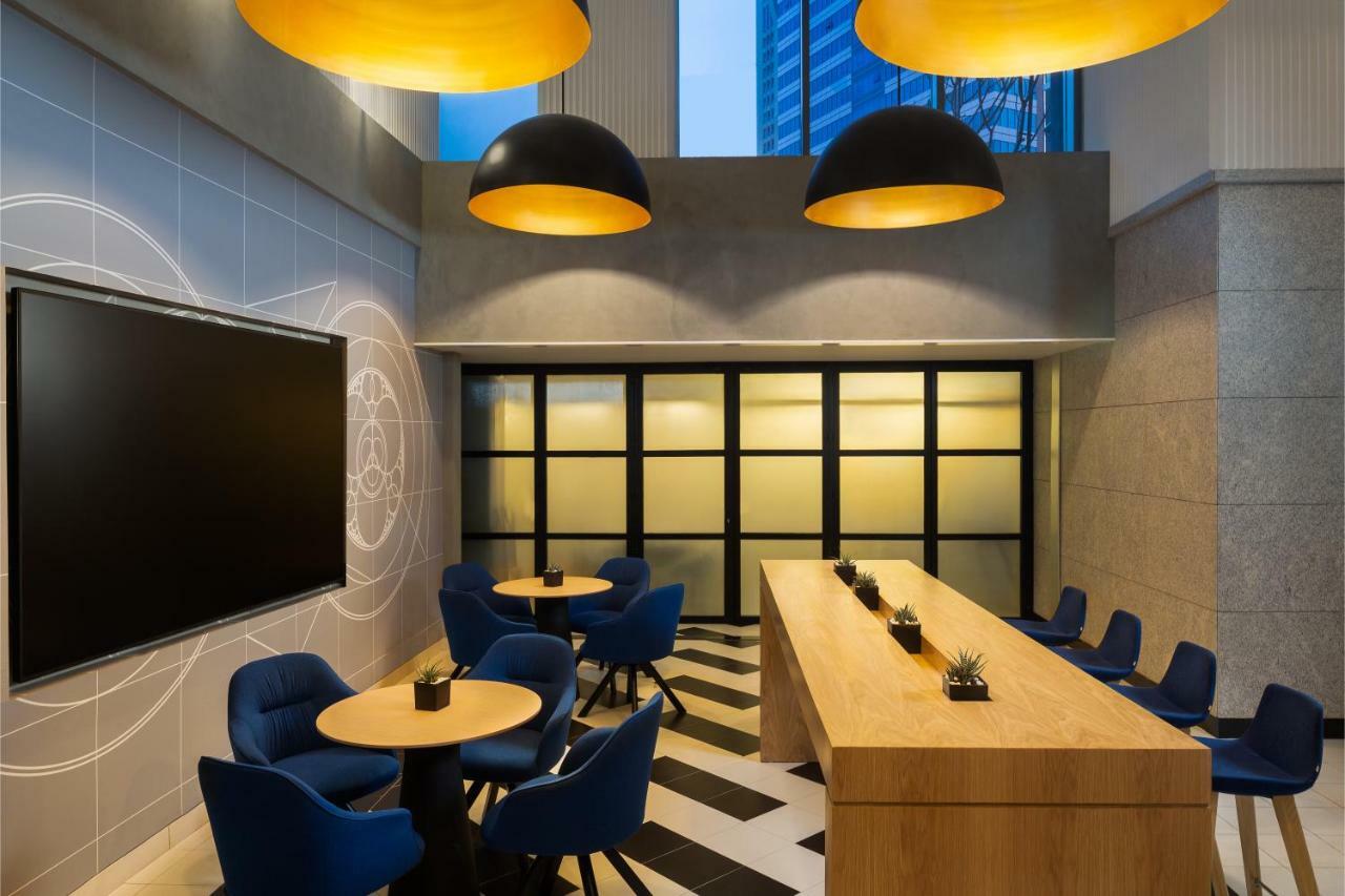 Millennium Atria Business Bay Aparthotel Dubaj Kültér fotó The photo shows a modern interior space, likely a lounge or meeting area. It features a long wooden table with some small plants on it, accompanied by several blue, circular chairs arranged around it. There is a large flat-screen television mounted o