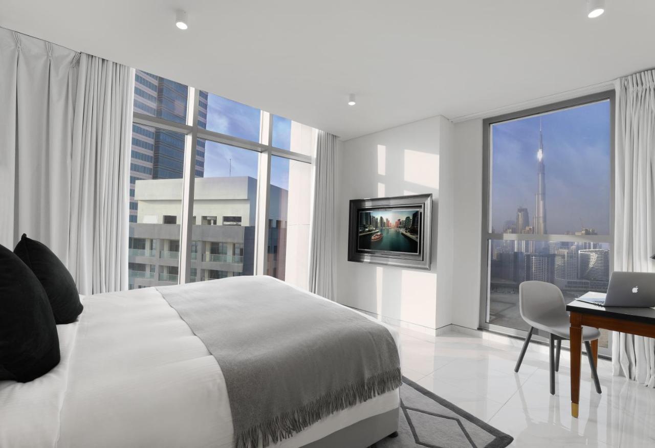 Millennium Atria Business Bay Aparthotel Dubaj Kültér fotó The photo depicts a modern hotel room that features a large bed with a gray throw blanket. There are large windows allowing natural light to fill the space, showcasing a city skyline view. A flat-screen television is mounted on the wall opposite the 